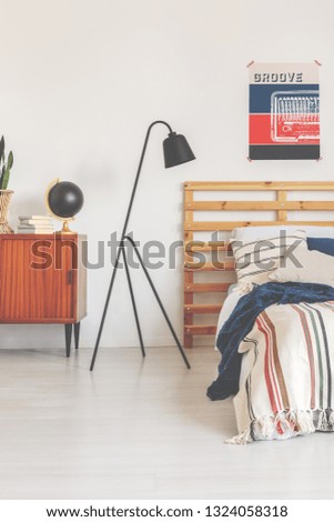 Real photo of a modern lamp in a retro bedroom interior with a bed, poster and wooden cupboard