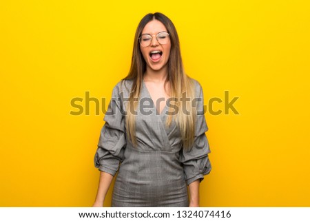 Young woman with glasses over yellow wall shouting to the front with mouth wide open