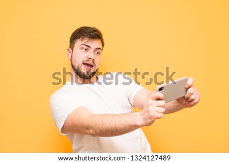 Happy adult man with a beard expressively playing video games on a smartphone on a yellow background. Joyful gamer in a white T-shirt plays mobile games on a colored background. Copyspace