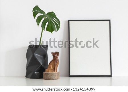 Black photo frame and origami flower pot with mostera leaf near white wall. Mock up