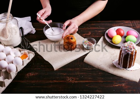 The girl prepares Easter baking, smears the cake with icing and sprinkles with colored powder. Preparing for holiday on the kitchen table, on a dark background.