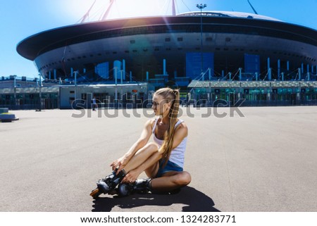 Portrait of a beautiful young blonde girl sitting on the ground asphalt and wearing roller skates, in denim shorts and a T-shirt. Hot summer day. Outdoor sports. Healthy lifestyle concept