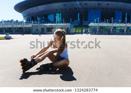 Portrait of a beautiful young blonde girl sitting on the ground asphalt and wearing roller skates, in denim shorts and a T-shirt. Hot summer day. Outdoor sports. Healthy lifestyle concept