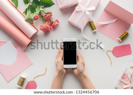 cropped view of woman holding smartphone near roses, rolls of paper, wrapped gifts, envelope, card and spools of thread on grey background