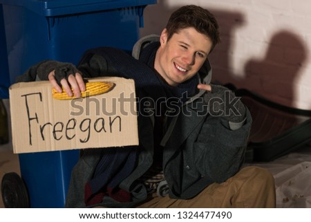 smiling homeless man holding corn cob and holding cardboard card with "freegan" inscription