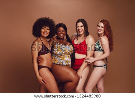 Group of women of different sizes wearing stylish bikinis and standing together against brown background. Diverse women in swimwear looking at camera and smiling.