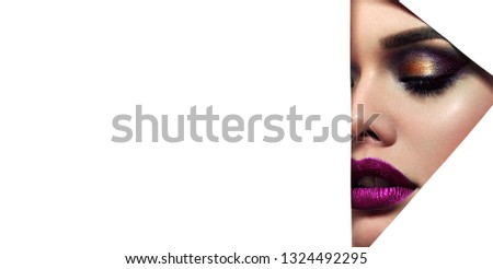 The face of a young beautiful girl with bright makeup and full purple lips peers into a triangular hole in white paper.