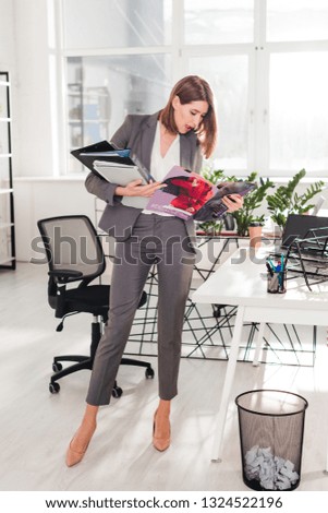 attractive businesswoman holding folders and reading magazine while standing in office 