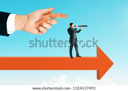 Side view of hand pointing at young businessman looking into the distance while standing on arrow. Sky background. Vision and search concept 