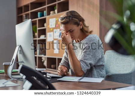 Exhausted businesswoman having a headache at office. Mature creative woman working at office desk feeling tired. Stressed casual business woman feeling eye pain while overworking on desktop computer. Royalty-Free Stock Photo #1324640339