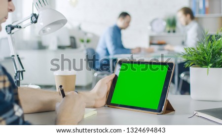 Close-Up of a Man's Hands Holding Green Screen Tablet Computer and Making Notes on Sticky Paper. In the Background Bright Modern Office with People Working in it.