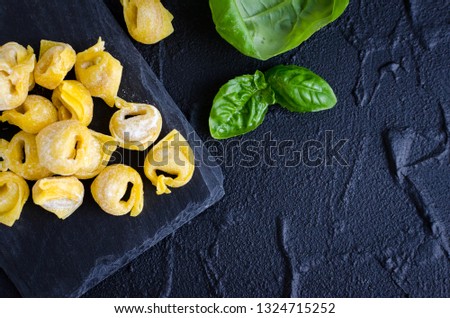 Freshly prepared homemade raw flouring tortellini ready to cook on dark stone plate with basil leaves on black background. Traditional Italian cuisine concept. Top view. Copy space.
