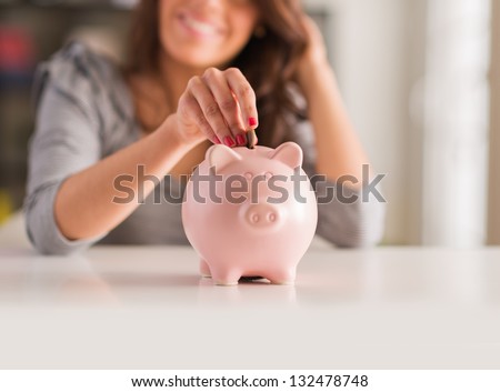 Woman Putting Coin In Piggy Bank, Indoors Royalty-Free Stock Photo #132478748