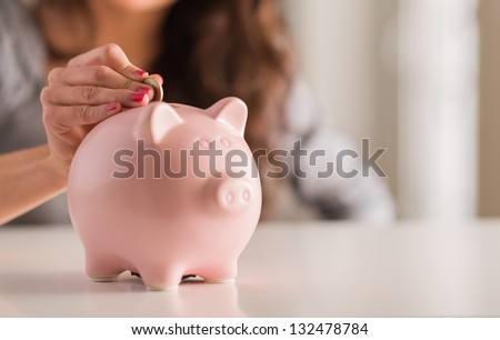 Woman Putting Coin In Piggy Bank, Indoors Royalty-Free Stock Photo #132478784