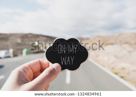 closeup of the hand of a young caucasian man holding a black signboard, in the shape of a thought bubble, with the text on my way written in it, on a road Royalty-Free Stock Photo #1324834961