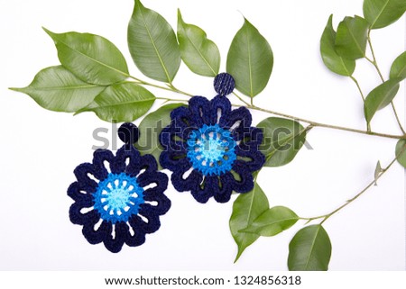 Closeup of a set of blue knitted flower shaped earrings on top of a branch and green leafs against a white background. Studio background shot.