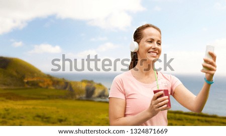 fitness, technology and healthy lifestyle concept - woman in earphones with takeaway smoothie or shake and smartphone listening to music after exercising over big sur coast of california background