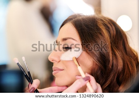Sophisticated beautiful caucasian brunette sitting in beauty salon while makeup artist putting blush on woman's cheeks.