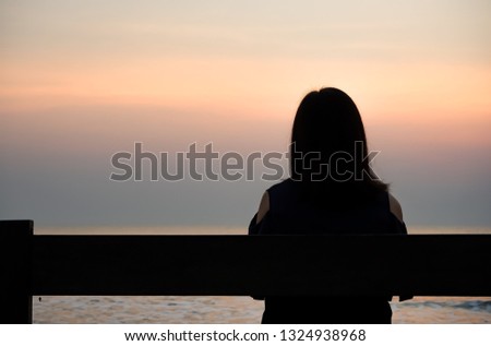 Back view silhouette of young girl sitting on wooden bench near the beach at sunset time. Concept of retirement, Relationship and divorce, Alone, Depression and sorrow, Hope, homeless, Peaceful. Royalty-Free Stock Photo #1324938968