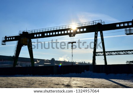 The sun's beam shines through a gantry crane with a hook for lifting and moving heavy loads. Construction site in the winter. Industrial plant
