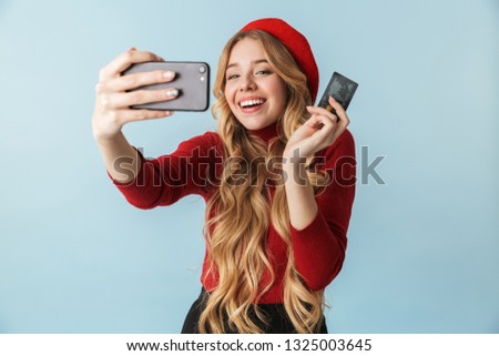 Attractive blond woman 20s holding credit card while taking selfie photo on cell phone isolated over blue background in studio