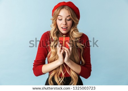 Image of surprised blond woman 20s wearing red beret holding present box isolated over blue background in studio