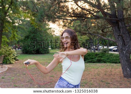 Young stylish cute girl is listening to music in headphones on mobile phone and dancing in the park. Pretty woman is relaxing and having fun.