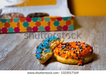 45 degrees picture of two donuts, blue and orange, over wood table and a box of donuts unfocused at the back