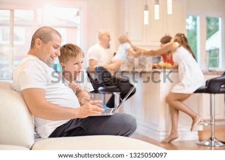 Son with his father sitting on the couch, looking at the laptop, in the kitchen mom friends and baby