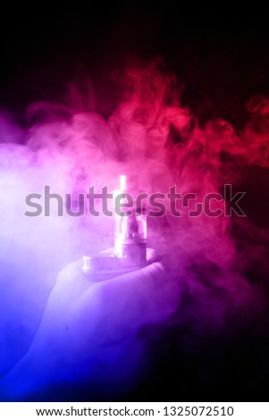 Male hand holds vape with atomiser in thick vape fog or smoke. Vape clouds around hand with vape on black background. Fog is dual color blue and red. Stock isolated colorful smoke with spray glycerine