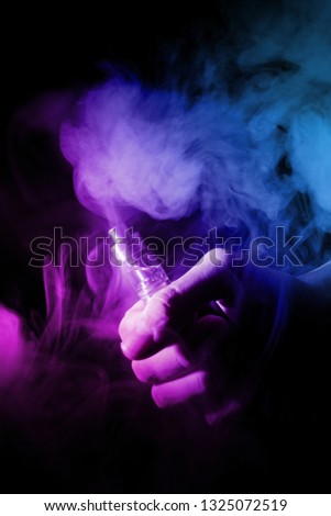 Male hand holds vape in thin vape fog or smoke. Vape clouds around hand with vape on black background. Light is dual color — blue and purple. Stock isolated colorful smoke with spray glycerine.