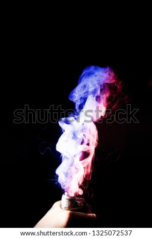 Male hand holds vape. Vape clouds splash from vape coil on black background. Fog is dual color — blue and red. Stock isolated dual colour smoke with spray boiling glycerine.