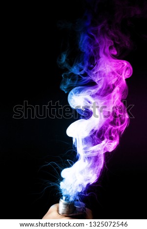 Male hand holds vape. Vape clouds splash from vape coil on black background. Fog is dual color — blue and purple. Stock isolated colorful smoke with spray boiling glycerine.