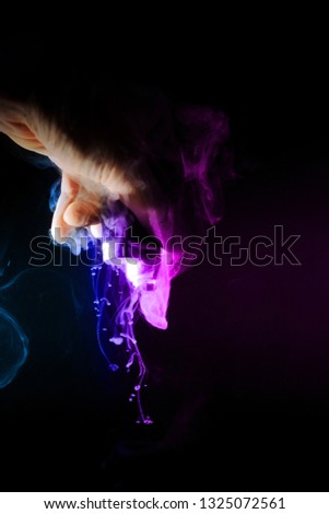 Male hand holds vape upside down. Vape clouds splash from coil on black background. Fog is two-colour blue and purple. Stock isolated dual color smoke with spray boiling glycerine.