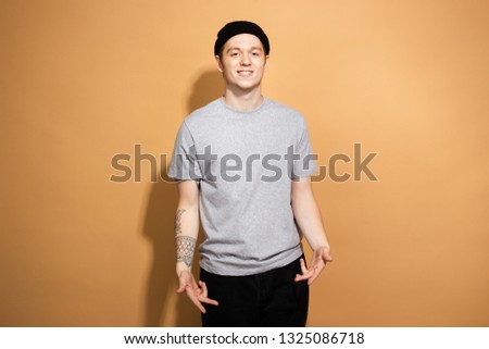 Smiling guy dressed in a gray shirt, black jeans and black hat with tattoo on his hand is posing on the beige background in the studio