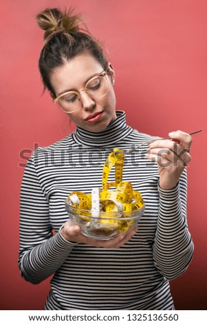 Shot of worried cute woman with eyeglasses holding bowl with measuring tapes over pink background.