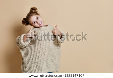 Happy young girl wearing beige huge sweater making thumb up sign and smiling cheerfully. Body language. I like that. Good job