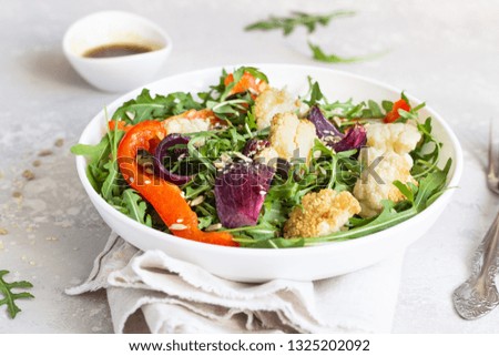 Salad with arugula, roasted vegetables (cauliflower, red bell pepper and onion) and seeds. Healthy food. Light grey concrete background. Copy space.