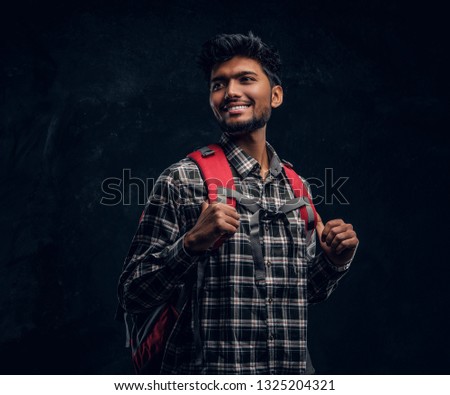 Portrait of a handsome Indian student with a backpack wearing a plaid shirt, smiling and looking sideways. Studio photo against a dark textured wall