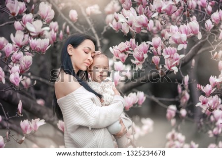Cute Baby 6 month old Girl in Pink Outfit with Big Blue Eyes with Young Beautiful Mother at Spring, Pink Blooming Tree at the Background.