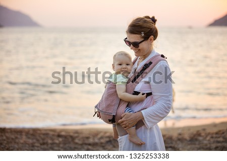 Cheerful Caucasian woman with baby daughter in buckle carrier on beach at sunset Royalty-Free Stock Photo #1325253338