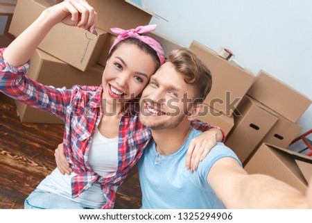 Young husband and wife moving to new place standing hugging taking selfie photos on smartphone looking camera smiling joyful holding key house purchase close-up