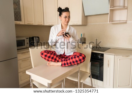 Brunette girl drinking black coffee at home in the kitchen. The girl sitting at the Desk in my pajamas. Bright kitchen. Pajamas. White shirt. Red plaid pants. Red bra.