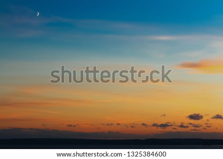 The sky is painted with bright colors from blue to orange and the moon complements the landscape.
