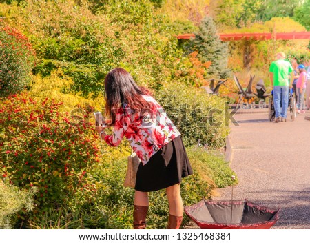 Woman with long dark hair taking photos of blooming bushes with her smartphone in park; people and autumn colors in background