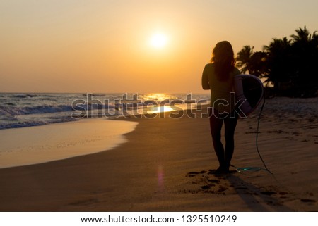 Woman surfer walking along the beach with a board in her hands against the sunset.