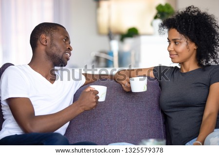 Satisfied African American couple talking, drinking coffee at home, smiling man and woman sitting on cozy sofa at home, having pleasant conversation, spending weekend together, resting Royalty-Free Stock Photo #1325579378