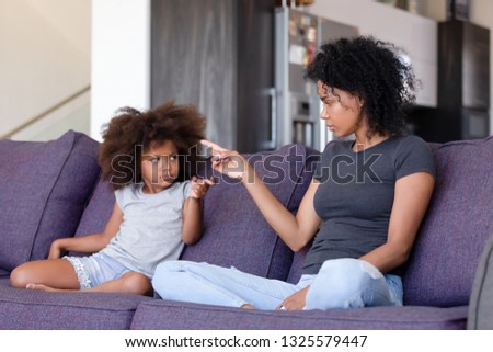 Upset African American woman and little girl quarreling at home, blaming each other, sitting together on sofa in living room, family conflict between offended mother and daughter