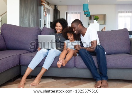 Happy African American family with daughter using laptop at home, shopping online, watching video or movie, smiling mother and father spending weekend together with child, sitting on sofa
