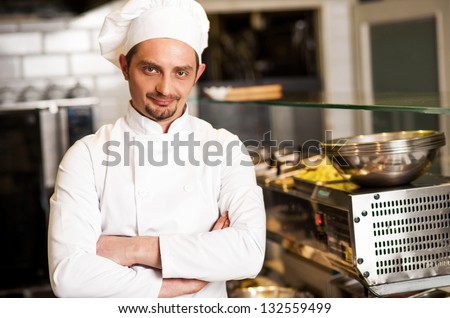Confident young male chef posing with arms crossed. Kitchen background.